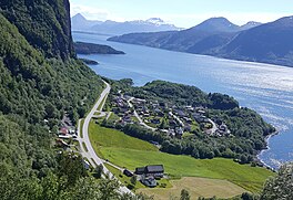 A small Norwegian village is depicted. We look down on it, surrounded by trees and other greenery. The village sits on the coast of a river, with islands on the backround. Parallel to the village is a road. A cliff face sits to the left of the road.