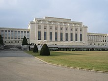 A drive leads past a manicured lawn to large white rectangular building with columns on it facade. Two wings of the building are set back from the middle section.