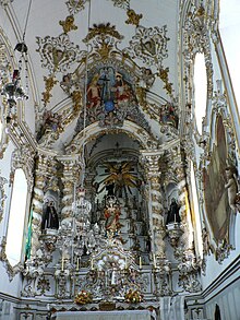 Photographie en couleurs. Dans un intérieur d'église très éclairé, un grand retable blanc avec des décorations dorées s'inscrit dans un cadre de la même configuration.