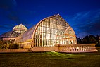 A one-story glass conservatory lit up yellow from the inside on a bluish night.