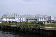 The outside of an association football stadium, with a river and foliage in the foreground.