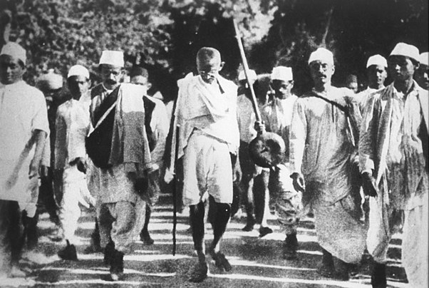 Old Black & White photo with central figure of Gandhi marching, in a loincloth, carrying a stick, with several followers marching alongside.