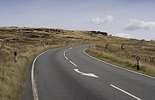 A flat, desolate, moorland under a cloudy sky, covered in long grass. A road runs from left to right in the distance.