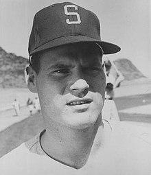 A man wearing a light baseball jersey and a dark cap with an "S" on the front