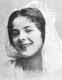 A smiling young white woman with dark center-parted hair with a white scarf draped over her head