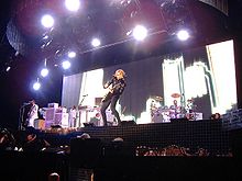 A female blond performer playing an electric guitar on stage. She is wearing black jacket and pants. The backdrops behind her display a numberof tall buildings. The stage is illuminated by a number of halogen lights from the ceiling.