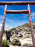 Photo couleur : vue en gros plan d'un torii, portique d'entrée d'un sanctuaire shintō, au sommet d'une montagne au sol parsemé de rochers, sous un ciel bleu nuageux.