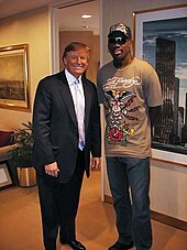 Donald Trump posing with basketball personality Dennis Rodman in a room with paintings adorning the walls. Trump is wearing a suit with a light-colored tie and dress shirt, while Rodman is wearing a brown t-shirt with a design on it, blue jeans, and a baseball cap that also has a design on it.