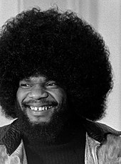 Photographie en noir et blanc d'un homme aux cheveux frisés, portant la barbe, souriant et regardant vers sa droite.