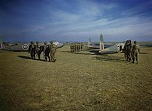 Three groups of six men wearing helmets and backpacks walk across a grass field towards waiting aircraft