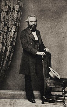 Photograph of Karl Marx standing over a chair, wearing a dinner jacket with neatly combed hair and beard