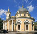 Seitliche Farbfotografie einer hellen, runden Kirche mit einer linken, kleinen Kuppel und einer rechten, großen Kuppel, die beide goldene Kugelspitzen haben. An beiden Eingängen befinden sich Säulen und Giebelfelder. Über den Fenstern sind kleine Rundbogenfenster mit Ornamenten.