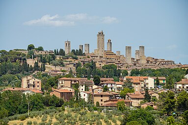 View of a small town on a hilltop surrounded by trees and vineyards. There are eight tall square towers rising from among the densely packed houses.