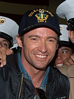 A man smiles for the camera as he wears a dark-colored jacket over a jean shirt and a cap possessing a navy emblem. He is surrounded by figures wearing white peaked caps, who are mostly cropped out of view.