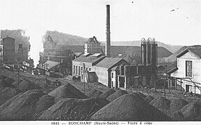 Photo noir et blanc montrant un complexe industriel avec bâtiments, trémies, cuves et cheminées entourés de terril, l'air est envahi de vapeurs et fumées.