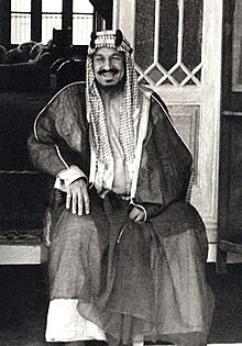 Un homme assis, souriant et moustachu porte une dishdasha sombre et un keffieh.