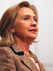 Informal pose of Clinton, in which she is wearing a beige wool jacket and facing toward the right. The angle at which Clinton is photographed is from her bottom right side.