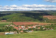 Village shown from the top of a hill. There are red and orange rooftops and green hills with cloud shadows in the distance.