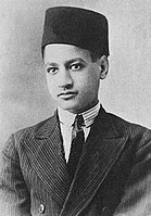 A boy wearing a jacket, a white shirt with a black tie and a fez on his head