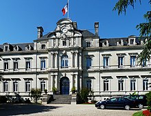 Photo de la façade principale ensoleillée du bâtiment de la préfecture qui s'élève sur trois étages imposants, couverts en ardoises. Au milieu, l'escalier d'une quinzaine de marches mène à un portail fermé à double vantail, entouré de chaque côté de deux colonnes. Au premier étage, autant de colonnes encadrent une grande fenêtre. Au-dessus, les colonnes ont fait place à des pilastres qui encadrent une petite fenêtre. Il est surmonté d'un fronton au milieu duquel se trouve une horloge. Sur le toit, un drapeau français flotte au sommet d'un mat.