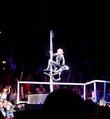 Faraway image of a stage and a woman hunched on a carousel horse which is attached around a pole. The woman wears a tight black dress with her hair in knots. Near the ground a long line of rods are visible, behind which the audience member are seen.