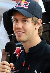 Sebastian Vettel mengenakan topi baseball bersponsor dan memegang mikrofon hitam di tangan kanannya