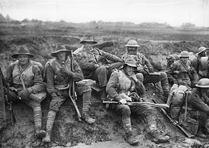 Black and white photo of six men wearing military uniform seated on a muddy slope in France, December 1916. Unidentified members of the Australian 5th Division, enjoying a smoko near Mametz, on the Somme. Some are wearing slouch hats, steel helmets, sheepskin jackets, and woollen gloves, demonstrating both the variety of official battledress, and how it was modified and augmented, for local conditions.