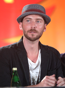 Photo en buste d'un homme avec les yeux bleus, portant une veste noire, un t-shirt blanc et un chapeau gris. Au premier plan se trouve une bouteille verte..