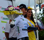 Photograhpie de trois-quarts d'un coureur cycliste, habillé en blanc, souriant, levant un bouquet de fleurs.