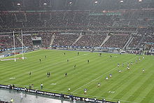 Vue depuis le haut des tribunes d'une rencontre de rugby. L'équipe à droite de l'image s'apprête à faire une remise en jeu, l'équipe opposée étant placée pour la réception.