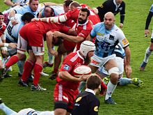 Un joueur de rugby court ballon en main en marge d'un regroupement d'autres joueurs des deux équipes.