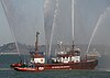 A red fireboat at rest on calm water sends five jets of water into the air