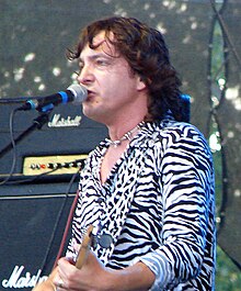 blurry image of Maťo Ďurinda a black-and-white zebra print shirt, playing electric guitar and singing into a microphone onstage