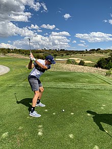 Photographie d'un homme en short gris, t-shirt blanc, vu de dos, jouant au golf, sous un ciel globalement ensoleillé