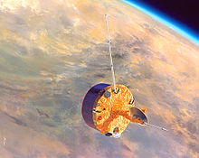 A stubby barrel-shaped spacecraft is depicted in orbit above Venus. A small dish antenna is located at the centre of one of its end faces