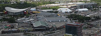 A downward-looking image of numerous buildings. A large arena is situated to the left, behind a building with a green peaked roof. To the right in the distance is a dirt race track.