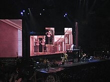 A female blond performer putting her right foot forward while standing on a stage. She wears a shiny corset and her hair falls around her in curls. There are black boots on her leg. The backdrop display a pink colored room with a French window and upholstery.