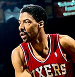 A basketball player, wearing a red jersey with the word "SIXERS" on the front, is looking to his right side.