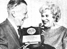 A white man and woman, both smiling, as he presents a plaque to her; the man is wearing a suit and holding the plaque with a hook-like prosthetic hand; the woman has coiffed light hair and is wearing drop earrings and a boatneck sweater