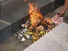 Pile of paper items on fire; a hand reaches to add money-like bills