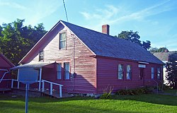 A 1+1⁄2-story wooden house, painted red, lit by the sun from the left with trees and part of a white house behind it.