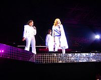 A blond woman wearing a white and purple leotard, with a cape around her, sings in to a microphone. She is flanked by two similarly dressed women.