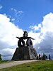 A path on green grass on a rise with white clouds and blue sky behind and a modern-looking sculpture in the background with some people next to it