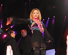 Picture of a middle-aged blond woman wearing a long-sleeve translucent black dress over black shorts and a top, the dress reaching down to mid-thigh. Brightly colored scarves are tied around her middle and shoulders. Around her neck she wears a number of colorful strings as necklaces. She stands with her hands stretched sidewards. Behind her, two men are seen facing each other.