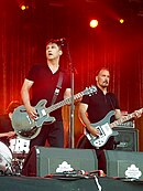 A picture of two men performing on stage, both wearing black shirts and playing electric guitars while being covered by red lights.