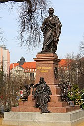 coloured photograph of a statue of a robed male figure on a stepped pedestal inscribed 'Felix Mendelssohn Bartholdy', with a seated female figure holding a lyre at its base, situated in an open space