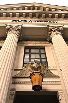 The building's entrance as seen from street level on Eighth Avenue. There is a metal lantern just above the door in the center. The door is flanked by triple-height marble columns that support a triangular pediment. The words "CVS Pharmacy" are mounted below the pediment.