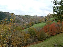 photo couleur d'une vallée encaissée. Des pâturages et zones boisées pentues à droite font face à des barres rocheuses abruptes à gauche.