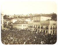 An old photograph showing a crowded square in front of a large, white, multi-storied building