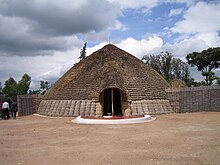 Foto istana Raja di Nyanza, Rwanda, menggambarkan pintu masuk utama, serta atap depan yang berbentuk kerucut.